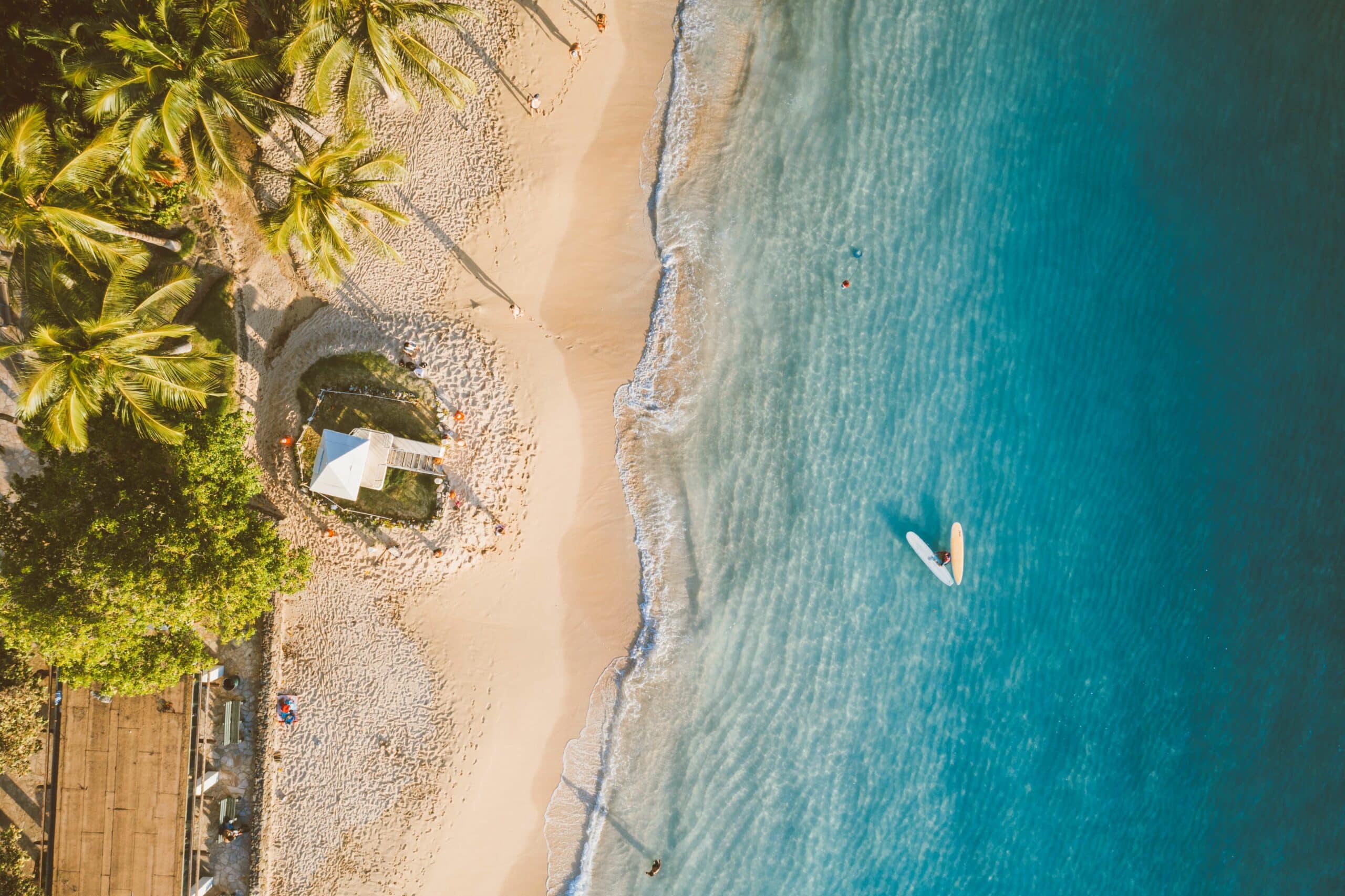 An aerial view of a white-sand beach with palm trees and a lifeguard station, with few people swimming.