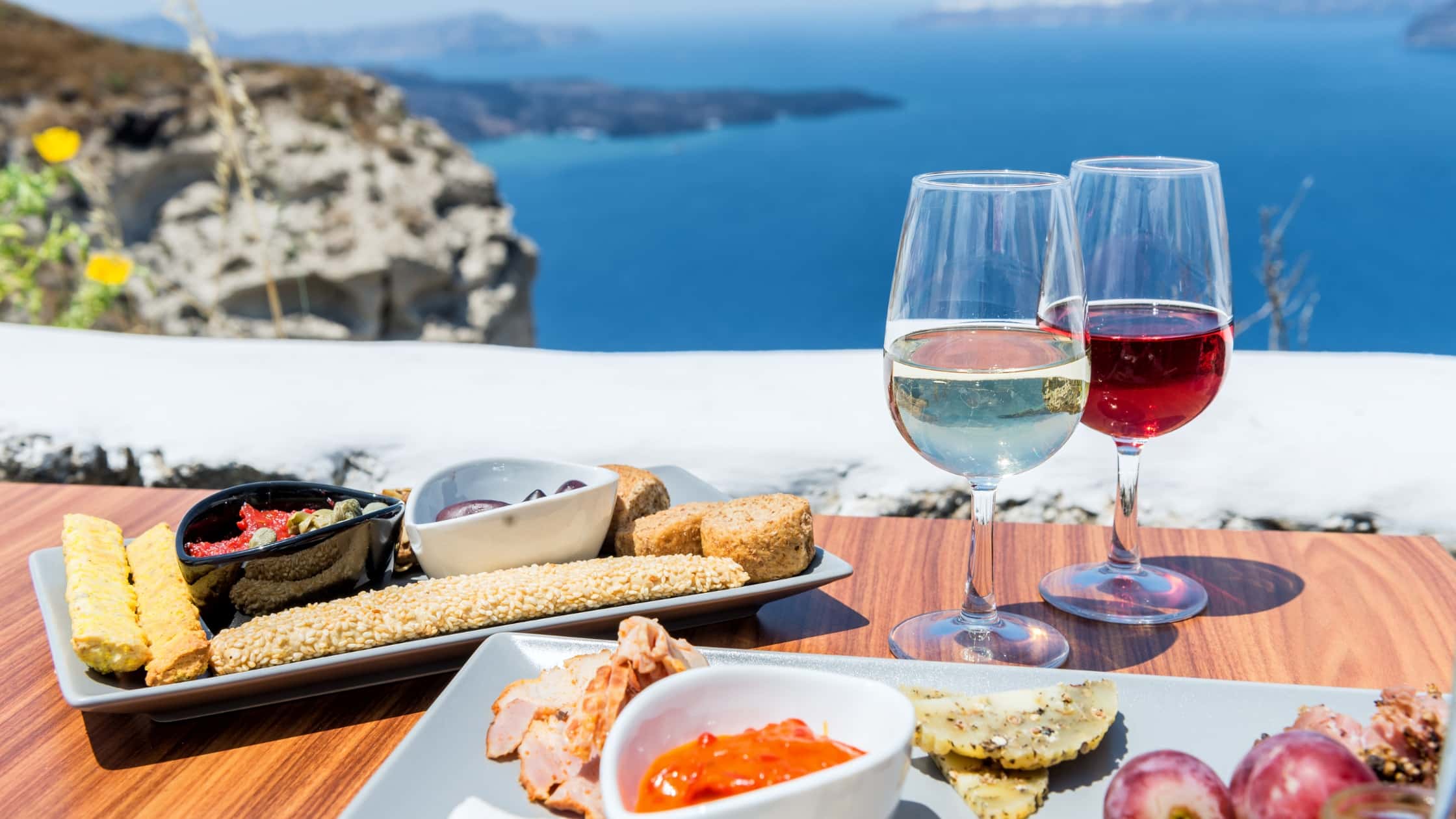 A closeup image of a charcuterie board with two glasses of wine with a background of the mountains and the sea during a wine tour in Greece.